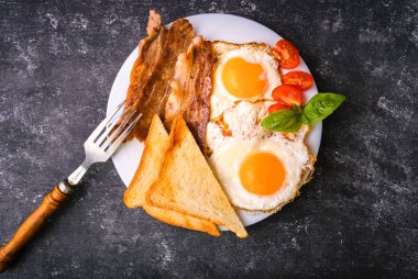 Pastırma ve kızarmış yumurta kahvaltı. Gevrek tost dilim dilimlenmiş kiraz domates, fesleğen yaprakları ile beyaz tabakta servis. Karanlık grunge beton tablo arka plan. Üstten görünüm, yukarıdan görünüm.