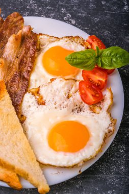 Pastırma ve kızarmış yumurta kahvaltı. Gevrek tost dilim dilimlenmiş kiraz domates, fesleğen yaprakları ile beyaz tabakta servis. Karanlık grunge beton tablo arka planı.