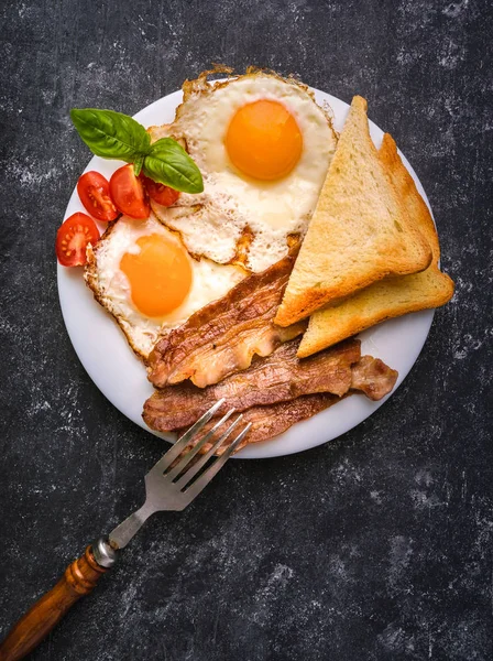 Pastırma ve kızarmış yumurta kahvaltı. Gevrek tost dilim dilimlenmiş kiraz domates, fesleğen yaprakları ile beyaz tabakta servis. Karanlık grunge beton tablo arka planı.