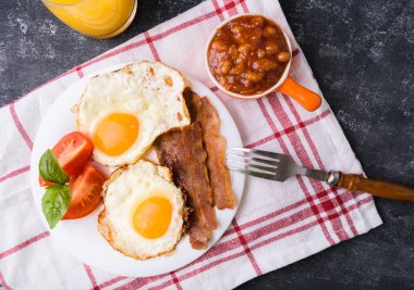 İngiliz kahvaltısı - pastırma, sahanda yumurta, fasulye ve portakal suyu. Dilimlenmiş domates ve fesleğen yaprakları ile beyaz tabakta servis. Karanlık grunge beton tablo arka plan. Üstten görünüm, yukarıdan görünüm.