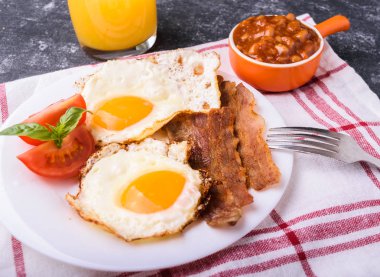 İngiliz kahvaltısı - pastırma, sahanda yumurta, fasulye ve portakal suyu. Dilimlenmiş domates ve fesleğen yaprakları ile beyaz tabakta servis. Karanlık grunge beton tablo arka planı.