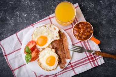 İngiliz kahvaltısı - pastırma, sahanda yumurta, fasulye ve portakal suyu. Dilimlenmiş domates ve fesleğen yaprakları ile beyaz tabakta servis. Karanlık grunge beton tablo arka plan. Üstten görünüm, yukarıdan görünüm.