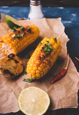 Izgarada mısır koçanı. Lezzetli yaz Aperatif. Maydanoz, pul biber, limon ve ızgara sarımsak. Eski ahşap kesme tahtası parşömen kağıt ile servis. Arka plan-koyu mavi taş beton.