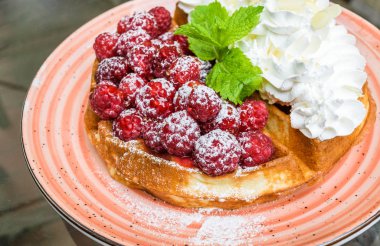 Taze ahududu ve badem pul ile krem şanti ile lezzetli Belçika Waffle. Nane yaprakları ile süslenmiş ve pudra şekeri, turuncu plaka üzerinde. Lezzetli yaz kahvaltı ya da tatlı.