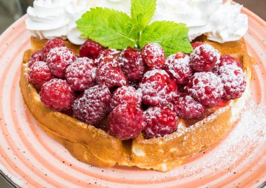 Taze ahududu ve badem pul ile krem şanti ile lezzetli Belçika Waffle. Nane yaprakları ile süslenmiş ve pudra şekeri, turuncu plaka üzerinde. Lezzetli yaz kahvaltı ya da tatlı.