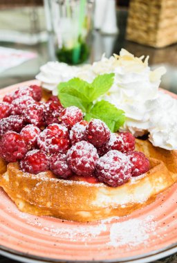 Taze ahududu ve badem pul ile krem şanti ile lezzetli Belçika Waffle. Nane yaprakları ile süslenmiş ve pudra şekeri, turuncu plaka üzerinde. Lezzetli yaz kahvaltı ya da tatlı.