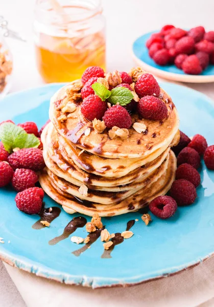 Ahududu ve çikolata şurubu, granola ile serpilir ve nane yaprakları ile süslenmiş ile Amerikan krep yığılmış. Taş masa lambası arka plan mavi tabakta servis. 