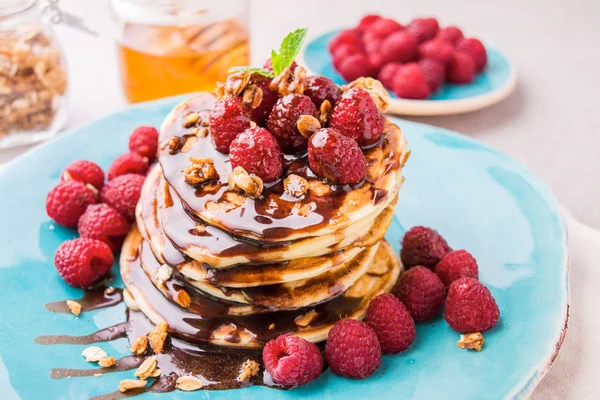 Ahududu ve çikolata şurubu, granola ile serpilir ve nane yaprakları ile süslenmiş ile Amerikan krep yığılmış. Taş masa lambası arka plan mavi tabakta servis. 