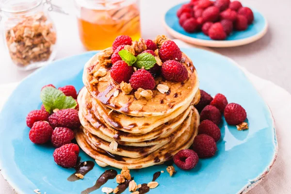 Ahududu ve çikolata şurubu, granola ile serpilir ve nane yaprakları ile süslenmiş ile Amerikan krep yığılmış. Taş masa lambası arka plan mavi tabakta servis. 