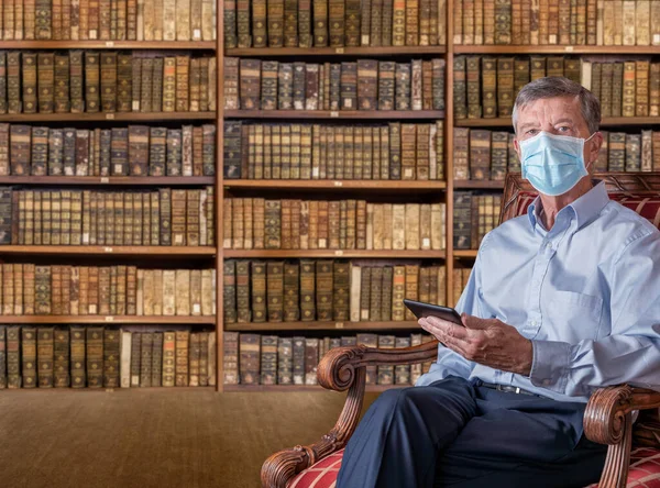 Bărbat în vârstă în mască de față împotriva cărții electronice de citire a virusurilor în biblioteca antică — Fotografie, imagine de stoc