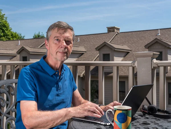 Starší muž pracující z domova na venkovní terase se sluchátky a počítačem — Stock fotografie