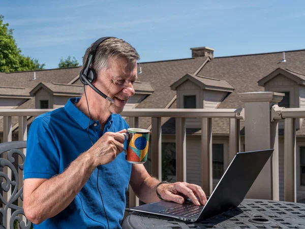 Homem sênior trabalhando em casa no deck ao ar livre com fone de ouvido e computador — Fotografia de Stock