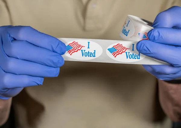 Gloved hands holding roll of I Voted stickers or buttons with USA flag ready for voter who voted in person in election — Stock Photo, Image