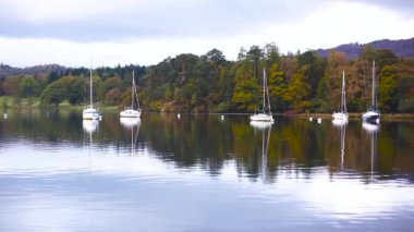 Yelkenli tekneler ve Lake District, İngiltere'de suda ağaçların yansımalar. Arka plan üzerinde bir woodland ile iskele yanındaki tekne demirlemiş. Sonbahar renkleri ve ruh hali, seyahat ve doğa kavramları