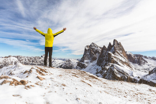 Successful man on top of snowy mountain. Happy hiker wearing a yellow jacket with raised arms looking at beautiful view on Italian Dolomites region. Adventure and travel concepts