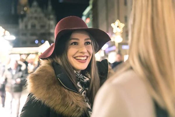 Happy women meeting and chatting at Christmas market in Munich. Brunette woman smiling at her blonde friend and looking at her. Christmas lights on background.