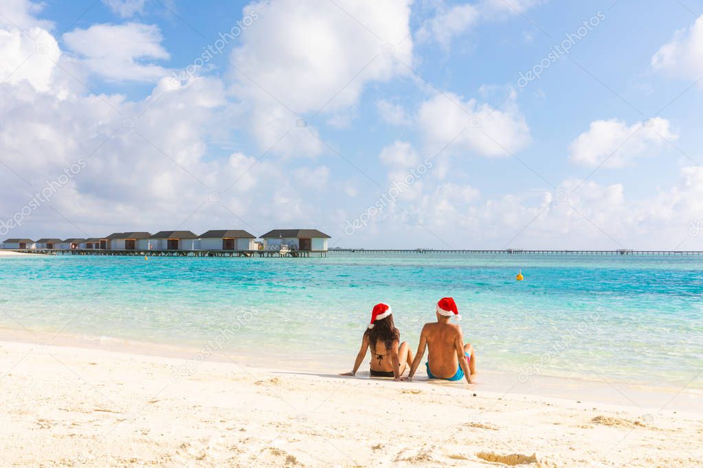 Couple with Christmas Santa hat relaxing at seaside. Holiday tropical destination postcard for holidays season with xmas hats. Man and woman sitting on the sand and looking away, travel and holidays concepts