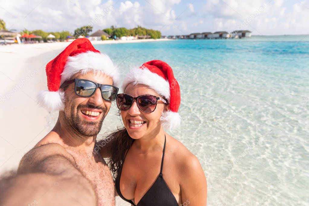 Smiling couple with Christmas Santa hat taking a selfie at seaside. Holiday tropical destination postcard for holidays season with xmas hats. Happy man and woman looking at camera.