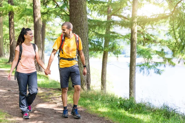 Happy couple walking in the wood holding hands. Young man and woman enjoying a summer day out together, walking on a lakeside path, looking each other and smiling. Love and healthy lifestyle