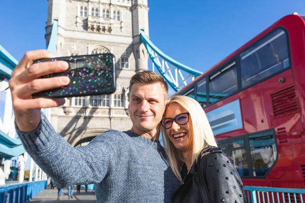 幸せなカップルの観光客がロンドンのタワー ブリッジで Selfie を取るします 男性と女性がスマート フォンを使ってセルフ ポートレートを取ると赤バスと背景にタワー ブリッジ 旅行とライフ スタイルの概念 — ストック写真