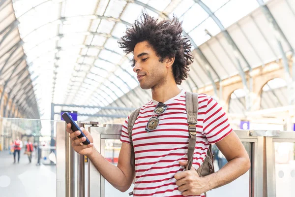 ロンドンの電車の駅で携帯電話を使用して幸せな男 巻き毛の笑顔と携帯電話に入力して混合レースの若い男 電車を待っています バックパッカー旅行とライフスタイル — ストック写真