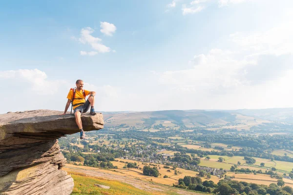 Man sitting on a rock cliff enjoying the view - Young man with backpack and hiking clothes on the cliffs over Hope Valley in the Peak District park, UK - Travel, nature and wanderlust concepts