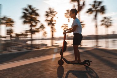 Gün batımında Malaga 'da e-scooter kullanan mutlu bir kadın - arka planda palmiye ağaçları ve seyahat fikirleriyle deniz kenarında yürüyüş yapan genç bir kadının yan görüntüsü - yaşam tarzı ve seyahat fikirleri