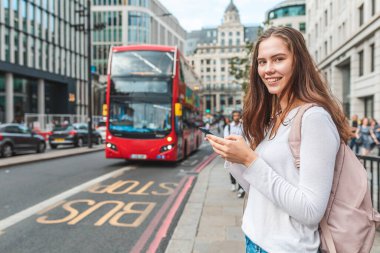 Londra 'da otobüs durağında akıllı telefonlu gülümseyen bir kadın - Londra' da bir gün otobüs tarifesini kontrol etmek için telefonunu kullanan gülümseyen bir kızın portresi - yaşam tarzı ve ulaşım kavramları