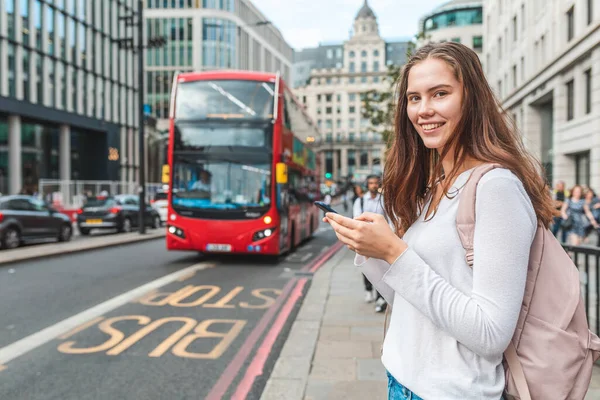 スマートフォンを持つ笑顔の女性がロンドンのバス停で ロンドンで1日のバスの時刻表をチェックするために彼女の携帯電話を使用して笑顔の女の子の肖像 ライフスタイルと交通概念 — ストック写真