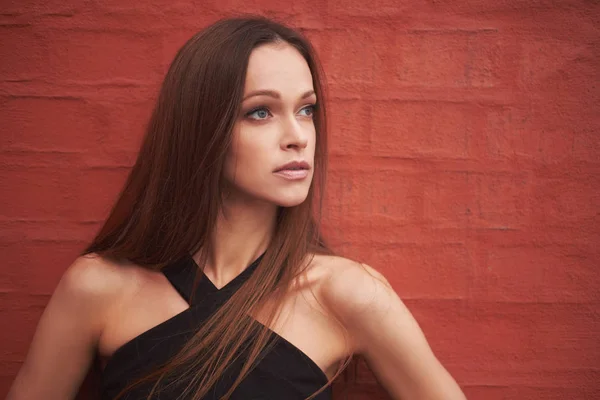 Young calm woman with long hair looking away standing on red brick wall background. Beautiful girl outdoors portrait