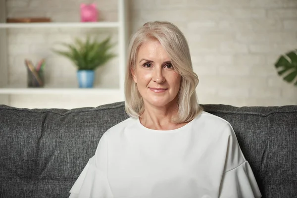 W średnim wieku blond kobieta siedzi na kanapie w domu — Zdjęcie stockowe