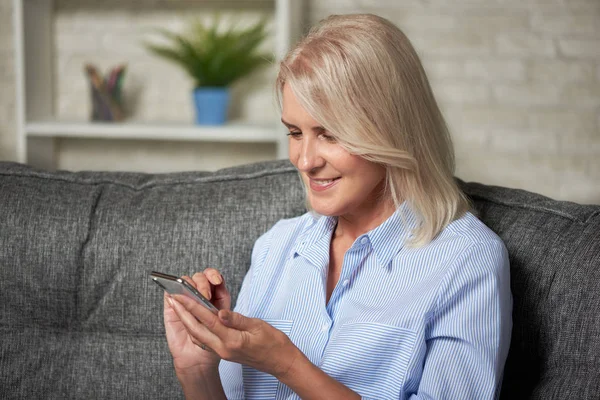 Starší usměvavá žena seděla doma na pohovce a držela se chytře — Stock fotografie