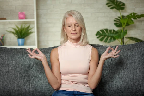 Hermosa mujer mayor sentada en un sofá y meditando en casa —  Fotos de Stock