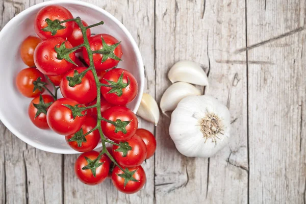 Fresh tomatoes in white bowl and raw garlic on rustic wooden tab — Stock Photo, Image