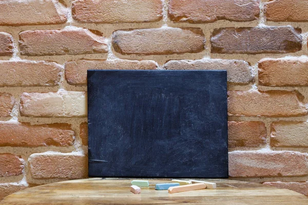 Blank blackboard on wooden table at brick wall.