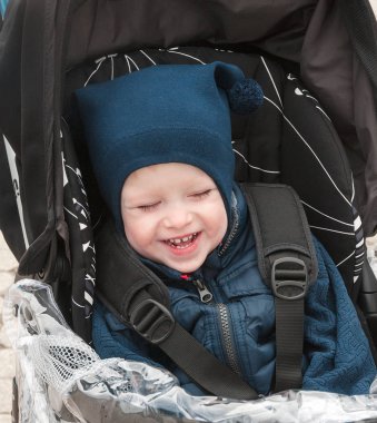 Sıcak mavi ceketli çocuk modern arabada oturur ve yürüyüşte gülüyor. Küçük çocuk. Çocuklarla seyahat. Bebek aile için ulaşım.