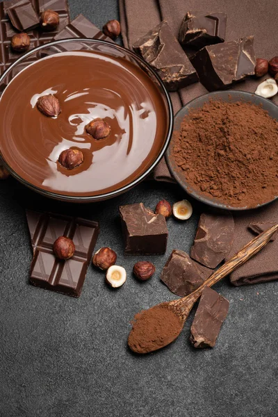 Skleněná miska z čokoládové smetany nebo roztavené čokolády, kousky čokolády a lískové ořechy na tmavém betonovém pozadí — Stock fotografie