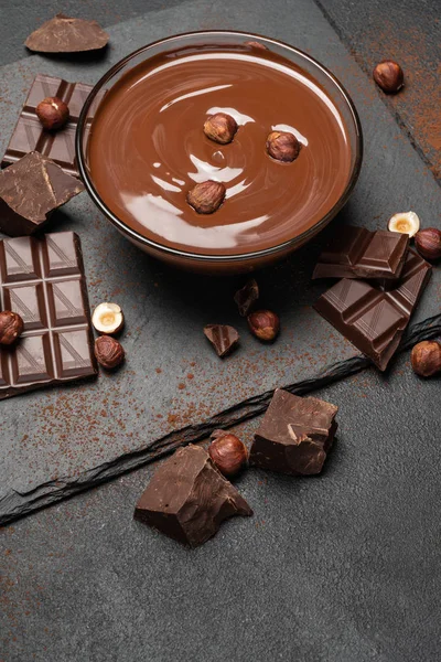 Semangkuk kaca krim coklat atau coklat cair, potongan coklat dan hazelnut di latar belakang beton gelap — Stok Foto