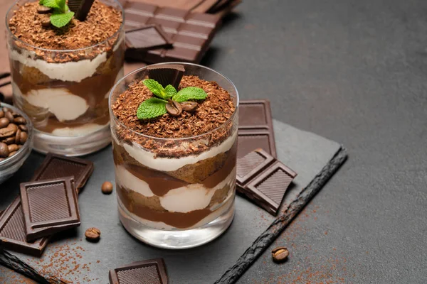 Sobremesa tiramisu clássico em um copo e pedaços de chocolate no fundo de concreto escuro — Fotografia de Stock