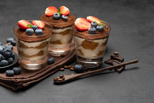 Makanan penutup tiramisu klasik dengan blueberry dan stroberi dalam gelas dengan latar belakang beton gelap — Stok Foto