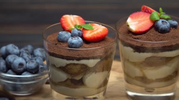 Classic tiramisu dessert with blueberries and strawberries in a glass on wooden background — Stock Video