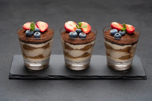 Classico dessert tiramisù con mirtilli e fragole in un bicchiere su tavola da portata in pietra su fondo di cemento scuro — Foto Stock