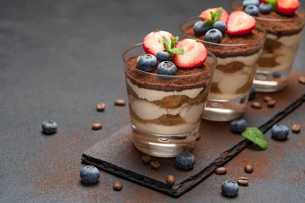 Klassisches Tiramisu-Dessert mit Blaubeeren und Erdbeeren im Glas auf Steinplatte auf dunklem Betongrund — Stockfoto