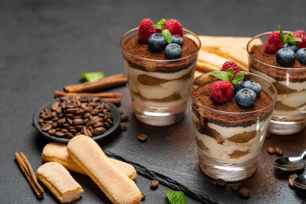 Sobremesa tiramisu clássica com mirtilos e framboesas em um copo e biscoitos savoiardi em placa de pedra no fundo de concreto escuro — Fotografia de Stock