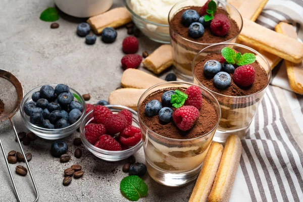 Sobremesa tiramisu clássica com mirtilos e framboesas em um copo e biscoitos savoiardi no fundo de concreto — Fotografia de Stock