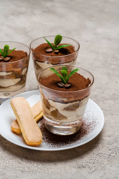 Sobremesa tiramisu clássico em uma xícara de vidro e biscoitos savoiardi na placa no fundo de concreto — Fotografia de Stock