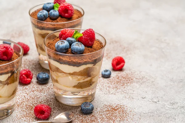 Klasyczny deser Tiramisu z jagodami i malinami w szklance i sitko z kakao w proszku na tle betonu — Zdjęcie stockowe