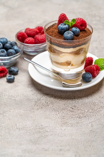 Klassiek Tiramisu-dessert met bosbessen en frambozen in een glas en kommen met bessen op een betonnen achtergrond — Stockfoto
