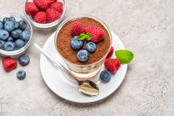 Klassiek Tiramisu-dessert met bosbessen en frambozen in een glas en kommen met bessen op een betonnen achtergrond — Stockfoto