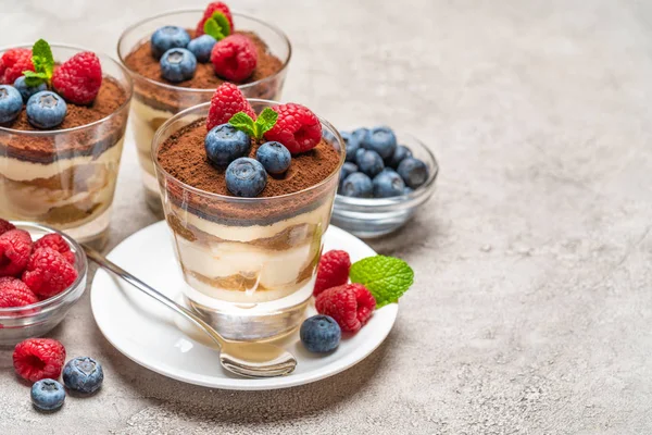 Sobremesa tiramisu clássica com mirtilos e framboesas em um copo e tigelas com bagas no fundo de concreto — Fotografia de Stock